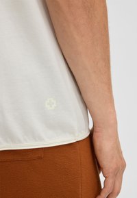 Kurzärmliges, hellbeiges T-Shirt mit einer weichen Textur, das ein dezentes Logo in der unteren linken Ecke trägt. Kombiniert mit braunen, strukturierten Shorts.