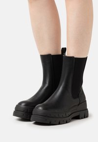 Schwarze Leder-Stiefeletten mit elastischen Seitenpaneelen, dicker Gummisohle, runder Zehenpartie und Zuglasche auf der Rückseite für einfaches Anziehen.
