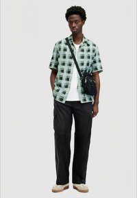 Camisa de manga curta em padrão xadrez nas cores verde menta e preto, combinada com calças cargo pretas e uma pequena bolsa preta de ombro. Modelo a usar sapatos bege.