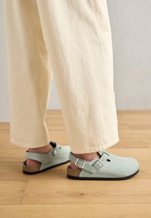 Stopy w jasnoniebieskich chodakach Birkenstock z dwoma srebrnymi klamrami, zestawione z kremowymi szerokimi spodniami na drewnianej podłodze.