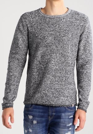 Mand iført en langærmet grå og sort meleret sweater kombineret med slidte blå jeans, stående foran en neutral baggrund.