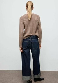 Beżowy sweter z prążkowanego dzianiny w połączeniu z szerokimi ciemnymi dżinsami, które mają podwinięte mankiety i tylne kieszenie. Całość dopełniają czarne buty.