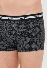 Boxer ajustados negros con un patrón geométrico en gris. Con una cinturilla elástica negra etiquetada como "HUGO" en blanco. Tejido suave y elástico.