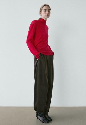 Frau steht, trägt einen leuchtend roten Rollkragenpullover, weite dunkelbraune Hosen und schwarze, mit Riemchen versehene Absatzsandalen vor einem einfarbigen hellen Hintergrund.