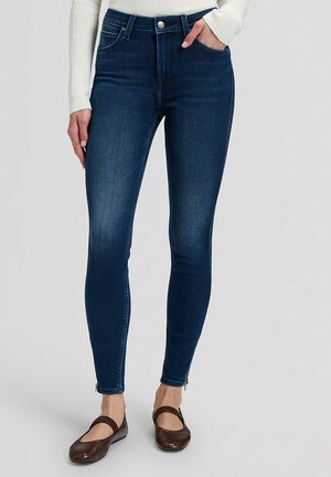 Donna che indossa jeans skinny blu scuro con cerniere alla caviglia, top bianco a coste manica lunga e ballerine Mary Jane marroni, in piedi su uno sfondo semplice.