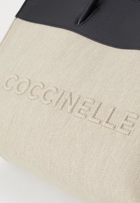 Borsa in tela beige con parte superiore in pelle nera. Presenta il nome del marchio in rilievo, "Coccinelle," sul davanti. Texture liscia.