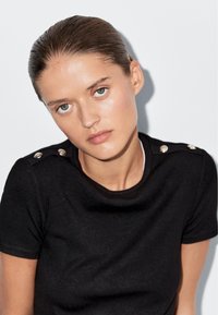 Zwarte korte mouwen top met een ronde halslijn en decoratieve knopen op de schouders, gemaakt van een gladde stof met een subtiele textuur.