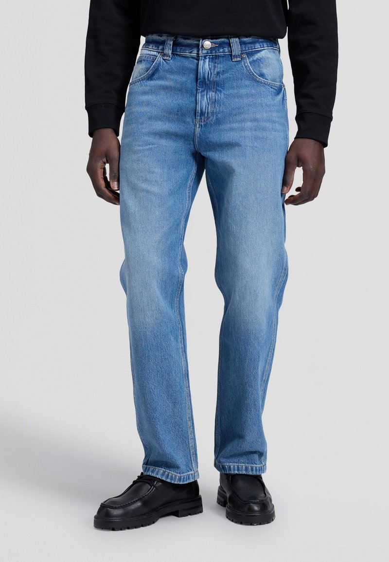 Homme portant un jean bleu clair droit, associé à des chaussures en cuir noires et un haut noir à manches longues, debout sur un fond uni.