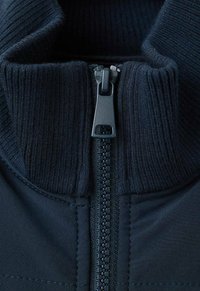 Zamaz blizu tamnoplave jakne s patentnim zatvaračem i rebrastim izrezom, prikazujući zupčanike i povlačenje patentnog zatvarača u jasnom detalju.