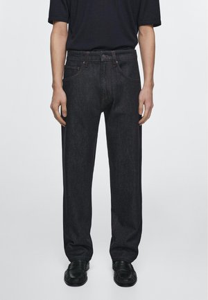 Persoon staat, draagt zwarte rechte jeans, zwarte loafers en een zwart T-shirt met korte mouwen tegen een effen lichte achtergrond.