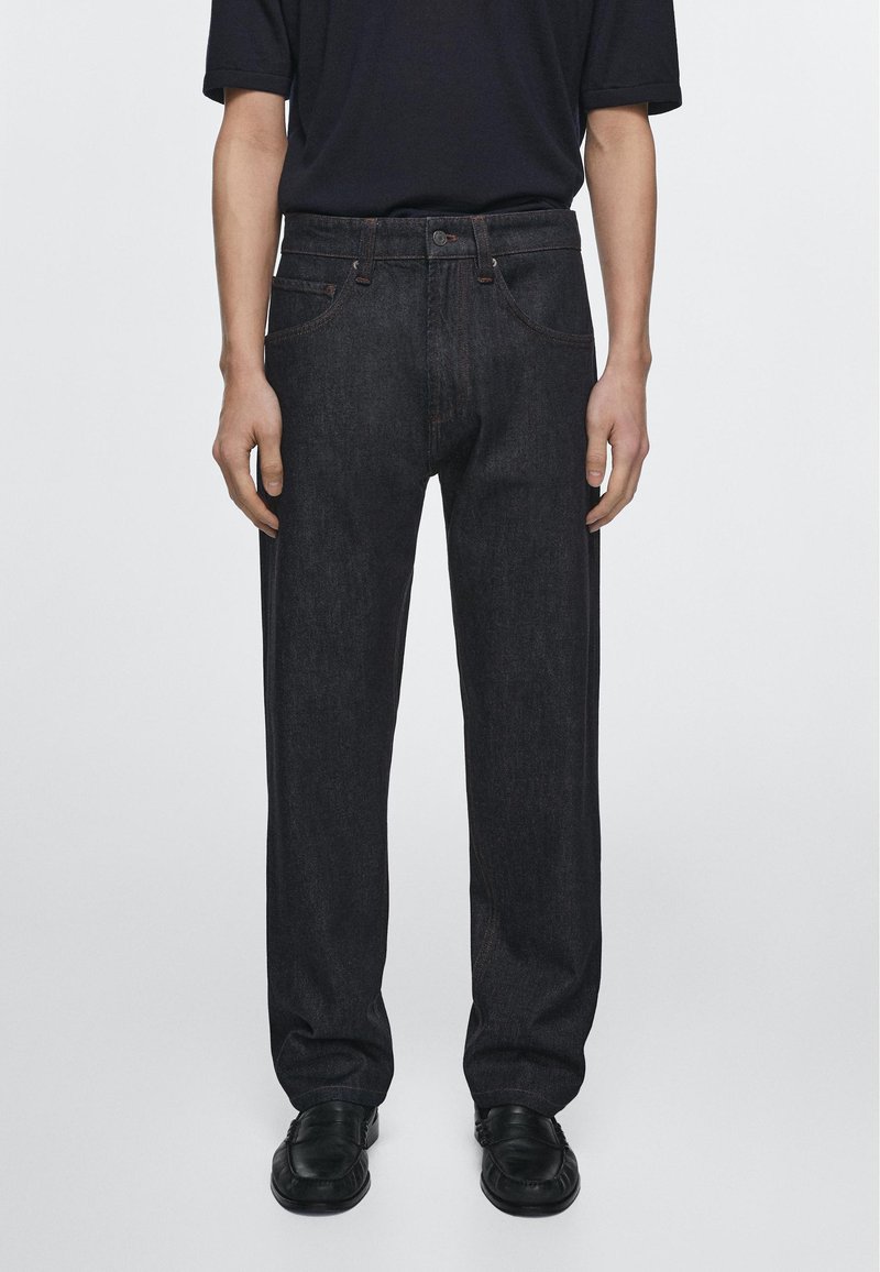 Person står iført sorte jeans med lige ben, sorte loafers og en kortærmet sort skjorte mod en ensfarvet lys baggrund.