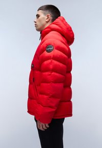 Chaqueta acolchada roja con capucha, con un acabado brillante, textura acolchada y un parche con logo en la manga izquierda. Bolsillo lateral con cremallera.