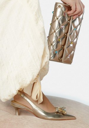 Mano sosteniendo un bolso de mano dorado acolchado con gemas coloridas, combinado con un zapato slingback dorado de tacón kitten con una gran decoración de cristal.