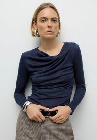 Mornarsko modra majica z dolgimi rokavi in drapirano ovratnikom, izdelana iz gladkega blaga. Model nosi srebrno zapestnico in vzorčast pas z karo hlačami.