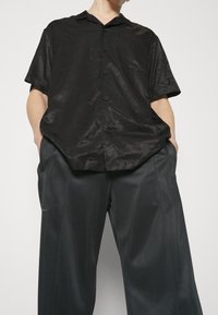 Musta lyhythihainen paita nappisuljennalla, jossa on lievää kiiltoa ja pehmeä pinta. Yhdistetty suurikokoisiin tummanharmaisiin housuihin.