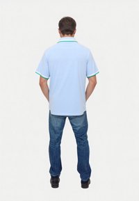 Hellblaues Poloshirt mit grünen Akzenten am Kragen und an den Ärmeln, kombiniert mit blauen Jeans und schwarzen Schuhen. Von hinten gesehen.