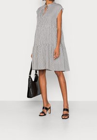 Zwart-witte gestreepte jurk met een gelaagd ontwerp, striksluiting bij de hals, korte mouwen, gecombineerd met zwarte schuursandalen en een gestructureerde handtas.