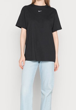 Personne portant un t-shirt noir Nike et un jean bleu clair, debout devant un fond clair uni.
