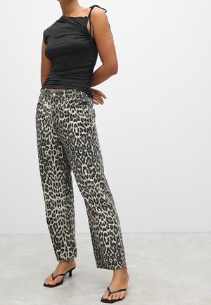 Woman wearing a black sleeveless top, leopard print wide-leg jeans, and black heeled sandals, standing with hands behind back.