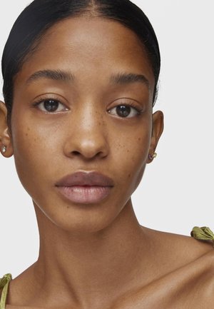 Primer plano de un rostro con un cutis liso y marrón claro, maquillaje natural y pequeños pendientes con piedras de colores. El cabello está peinado hacia atrás.
