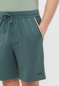 Calções verde-azulado escuros com cintura ajustável por cordão, bolsos laterais com detalhes em branco e dourado, e um pequeno logótipo preto "BOSS" na perna.