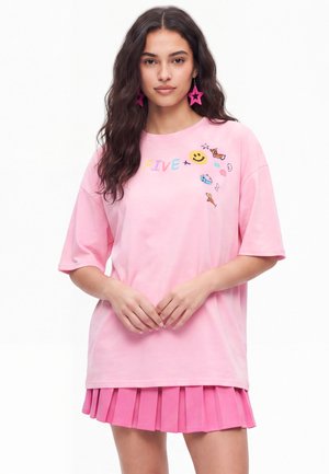 Kobieta o długich ciemnych włosach nosi różowy oversize T-shirt z kolorowymi bazgrołami i napisem „FIVE”, zestawiony z różową plisowaną spódnicą oraz kolczykami w kształcie gwiazdek.