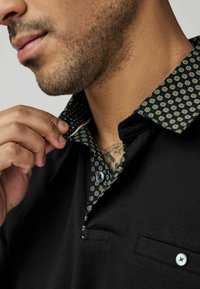 Černé polo tričko s vzorovaným límcem, který má geometrické vzory v greens a zlaté barvě. Viditelný jeden knoflík, zdůrazňující detailní šití.