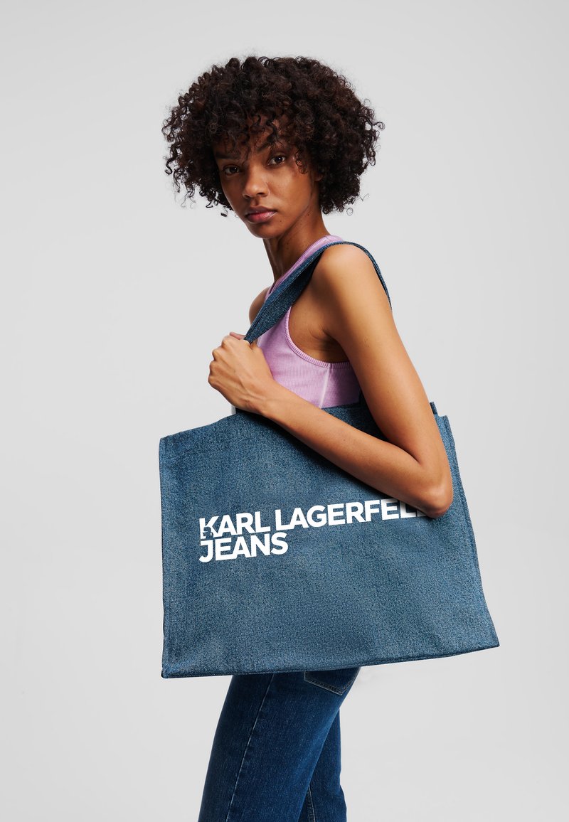 Tamsiai mėlynas džinsinis rankinis krepšys su tekstūruota apdaila. Vienoje pusėje yra dideli balti užrašai "KARL LAGERFELD JEANS". Reguliuojama petneša.