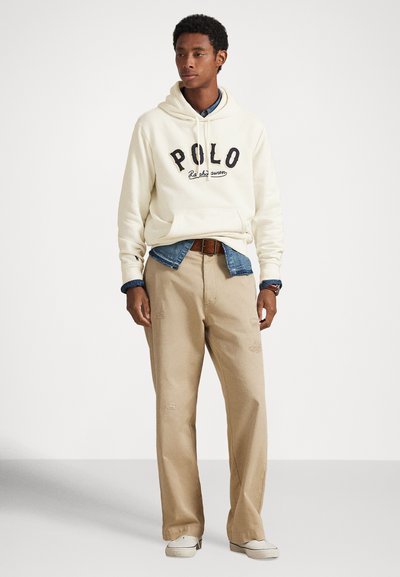 Jaunas vyras, vilkintis kreminės spalvos Polo džemperį su kapišonu virš džinsinio marškinio, smėlio spalvos kelnes su rudos spalvos diržu ir šviesios spalvos batus, stovintis atsipalaidavęs su rankomis žemyn.