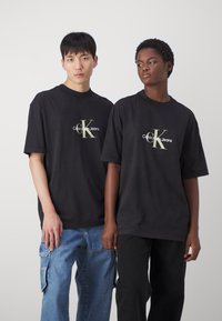 Černé oversized trička mají bílou a zlatou logo "Calvin Klein Jeans". Modely zahrnují volné džíny a tmavé kalhoty.