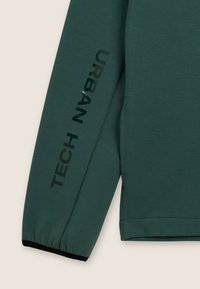 Haut à manches longues vert foncé avec des poignets noirs, présentant des inscriptions en lettres noires « TECH » et « URBAN » imprimées le long de la manche. Texture lisse.