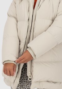 Des mains ajustant le poignet d'une doudoune beige clair sur des vêtements superposés, avec un pantalon à imprimé léopard visible.