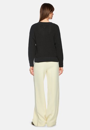 Woman wearing a black textured sweater and cream wide-leg pants, standing facing away against a plain background.