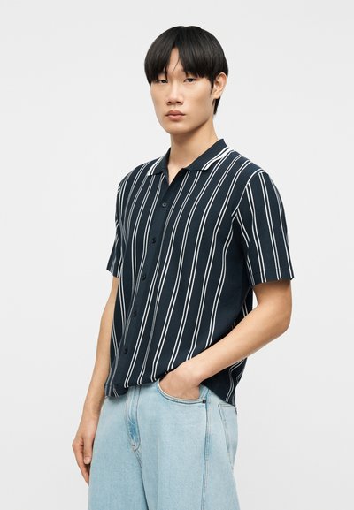 Νεαρός με ίσια μαύρα μαλλιά, που φοράει κοντομάνικο μαύρο πουκάμισο με λευκές κάθετες ρίγες και ανοιχτό μπλε τζιν, όρθιος με το ένα χέρι στην τσέπη.