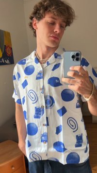 Camisa de manga curta com botão em tecido branco, apresentando padrões gráficos em azul de vários itens, incluindo chapéus, discos e caixas.