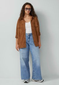 Bruine suède knoopjesoverhemd over een witte tanktop, gecombineerd met lichtblauwe wijdvallende jeans en grijze sneakers. Overmaatse zonnebril.