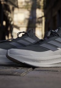 Ζευγάρι γκρι και μαύρα αθλητικά παπούτσια Adidas σε λιθόστρωτο δρόμο με θολά κτίρια της πόλης στο φόντο.
