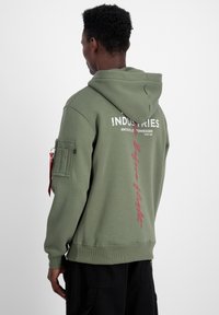 Hanorac cu glugă de culoare verde măslin, cu grafica text alb și roșu, buzunar frontal și un buzunar cu fermoar pe lateral. Material moale cu manșete ribbate.