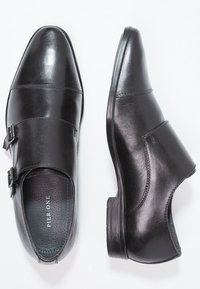 Des chaussures de ville pour hommes en cuir noir avec des boucles à double bride, une vue de haut de l'une des chaussures et une vue de côté de l'autre sur un fond blanc.