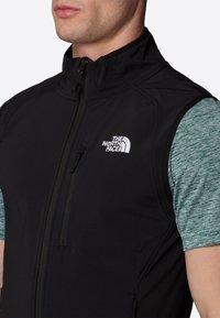 Black sleeveless vest with a high collar, zip closure, and side pocket. Features a white logo on the left chest and a textured blue shirt underneath.