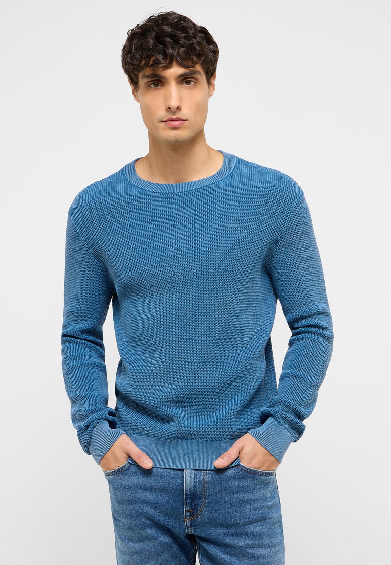 Young man with short curly hair wearing a blue knit sweater and blue jeans, standing with hands in pockets against a plain background.