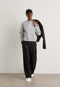 Gray sweatshirt with logo, black wide-leg pants, and white sneakers. Black cap worn forward and leather jacket draped over one arm.