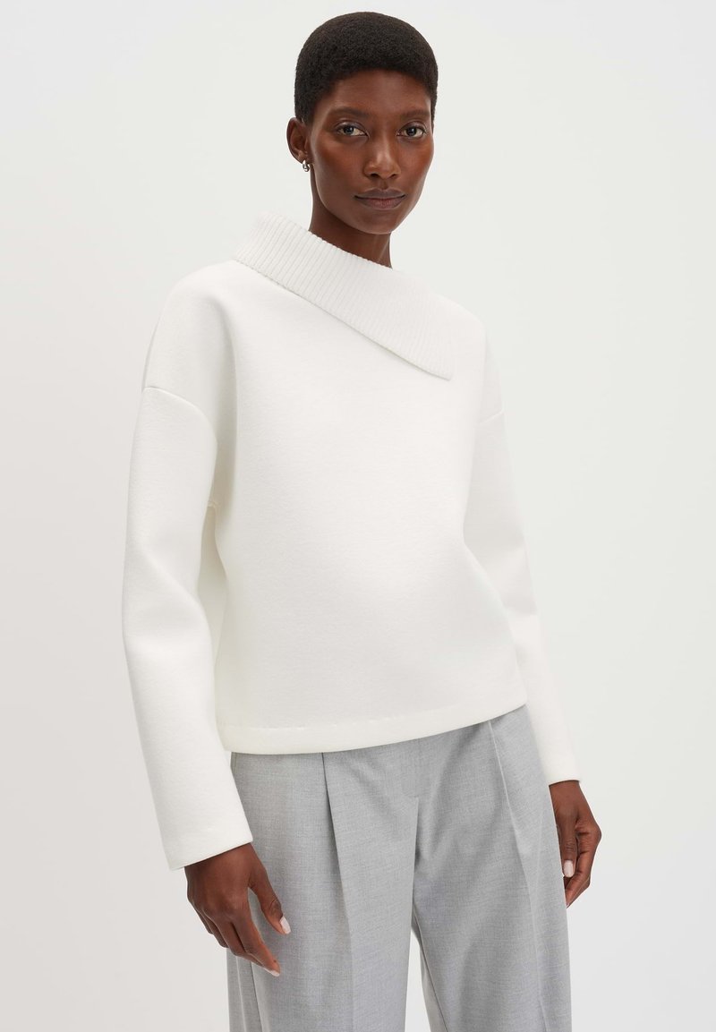 Pullover bianco strutturato con colletto asimmetrico, maniche lunghe e vestibilità rilassata, abbinato a pantaloni tailored grigi chiari.