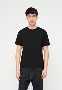 Νεαρός άνδρας που στέκεται και κοιτάζει μπροστά φορώντας μια απλή μαύρη κοντομάνικη μπλούζα και σκούρο τζιν, μπροστά από έναν απλό λευκό φόντο.