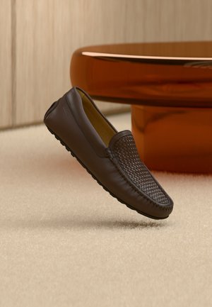 Mocassino in pelle marrone con dettaglio intrecciato sulla parte superiore, posizionato in verticale su un tappeto beige vicino a una base tavolo rotonda di colore ambra.