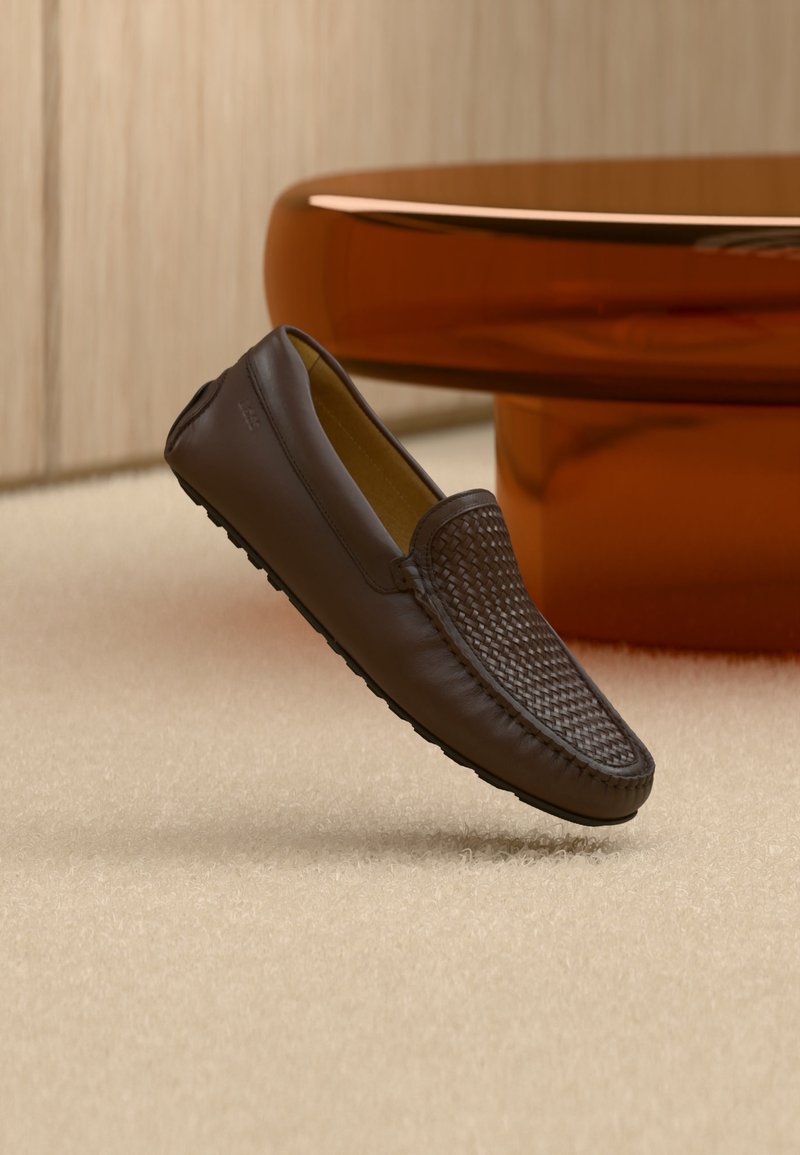 Mocassin en cuir marron avec un détail tissé sur le dessus, placé debout sur un tapis beige près d'une base de table arrondie de couleur ambre.