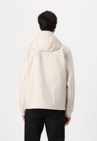 Light beige jacket with a hood, elastic cuffs, and a slightly textured fabric. Features a red accent along the neckline.