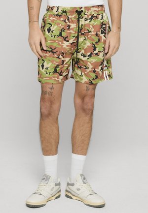 Mężczyzna ubrany w zielone, pomarańczowe i czarne moro szorty z czarnym sznurkiem, białe skarpetki i białe sneakersy New Balance, stoi na szarym tle.