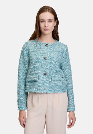Veste texturée turquoise avec trois boutons, encolure ronde et deux poches avant. Portée par-dessus un pantalon de couleur claire.
