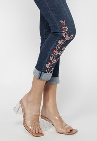 Jeans de mezclilla azul oscuro con puños enrollados, adornados con bordados florales rojos a lo largo del costado. Combinados con sandalias de tacón transparente.